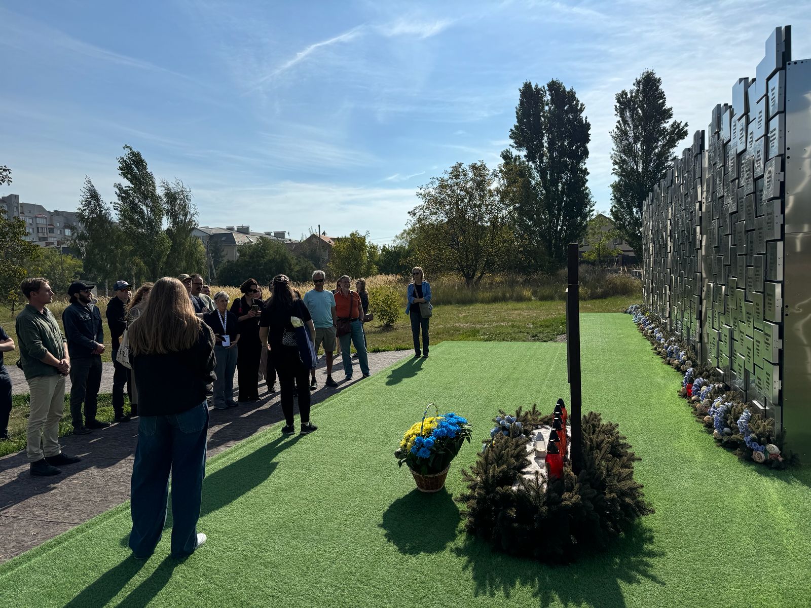 The group stands before a memorial wall of names and wreaths